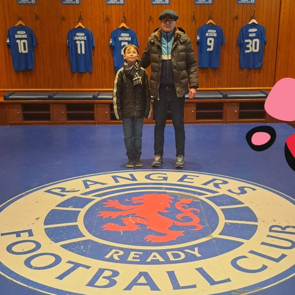son and dad standing in the rangers glasgow football changing room infront of the shirts